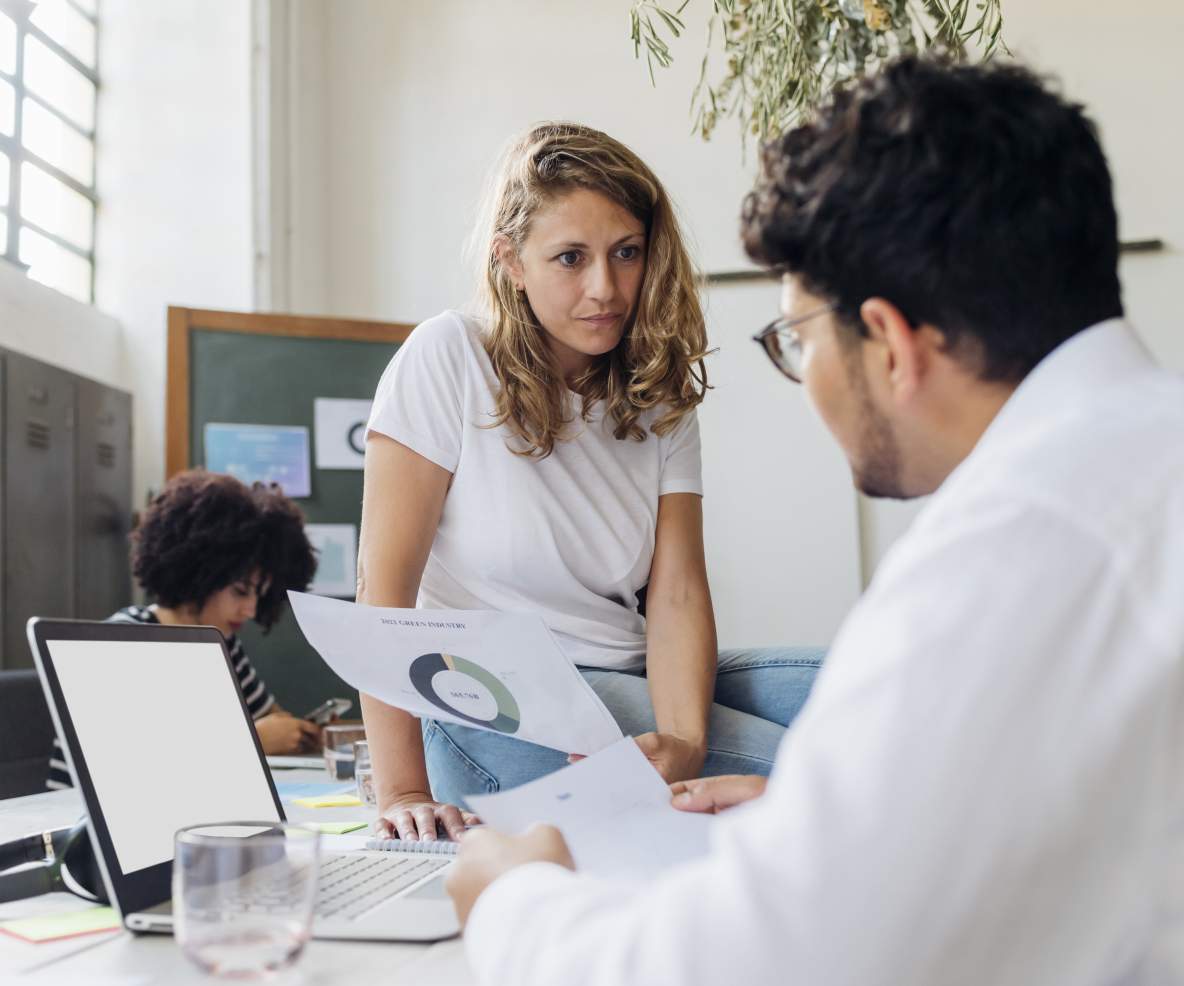 Vrouw zit op een bureau en luistert naar een mannelijke collega die aan het bureau zit